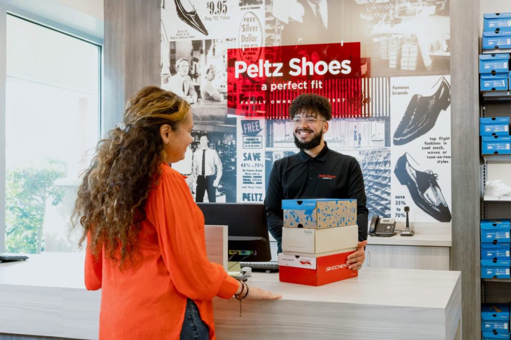 Peltz Shoes sign behind young man sales associate. 3 shoes boxes on a counter next to cash register. Woman in orange shirt at register checking out.