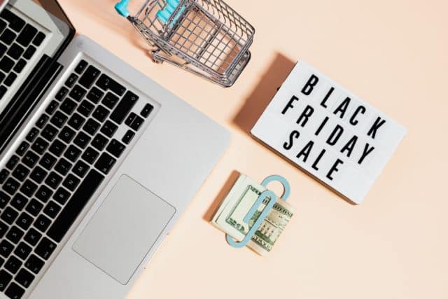 black friday sale sign; laptop; money in money clip; small toy cart on pink table top