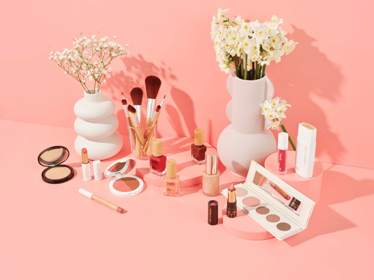 nourished life cosmetics on pink background; includes makeup brushes in a clear cup, eye shadow palette, face powder, nail polish, lip gloss, foundation, lip liner, and bronzer and blush duo. Sitting amongst the cosmetics are white flowers in white vases.