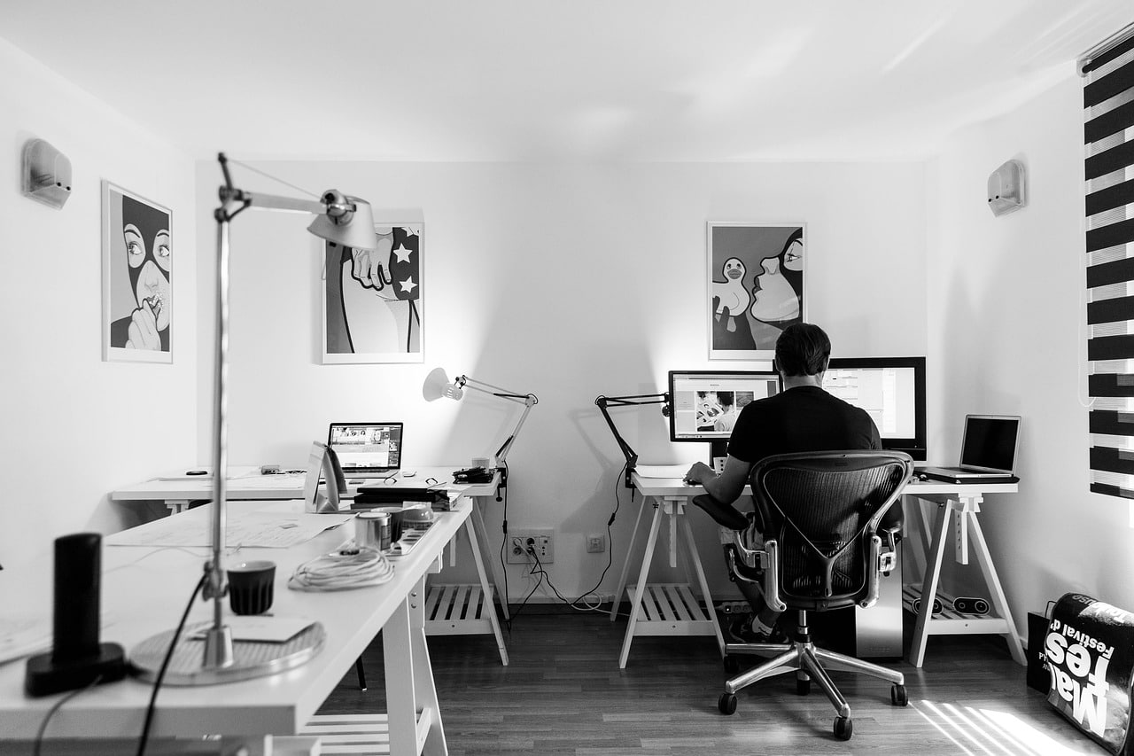 man sitting at computer coding; black and white image with drafting table on one wall and silver lamp; computer in the corner; 3 pieces of wall art