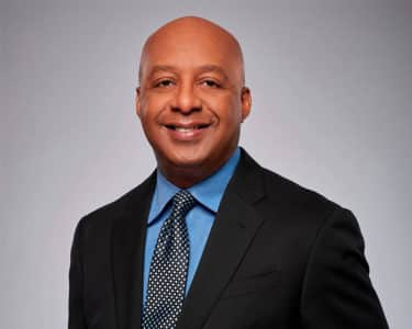 marvin ellison headshot African American man _ CEO headshot _ bald, smiling Black man in black suit with blue button up and tie in front of a grey backdrop
