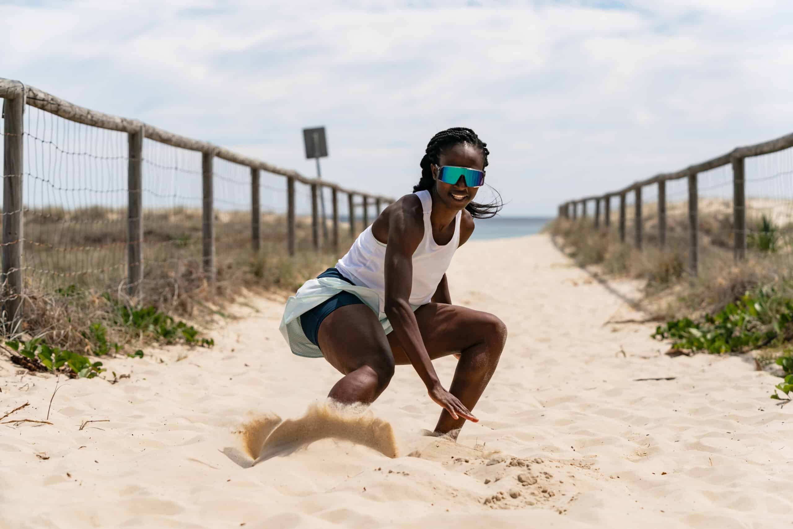 2xu image Black woman athlete in spandex shorts and white tank top and sunglasses , close to the ground in the sand with wooden railings on either side