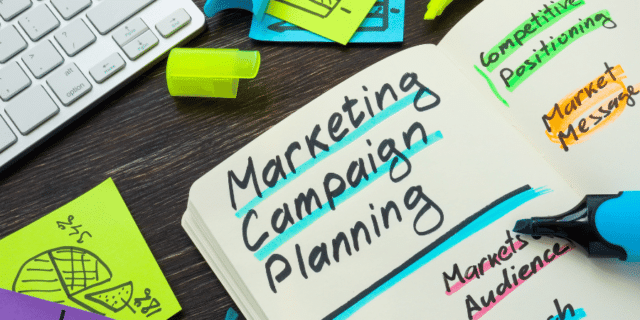 "marketing campaign planning" written in notebook - black marker with blue highlighter bright yellow sticky note and keyboard on dark wood desk